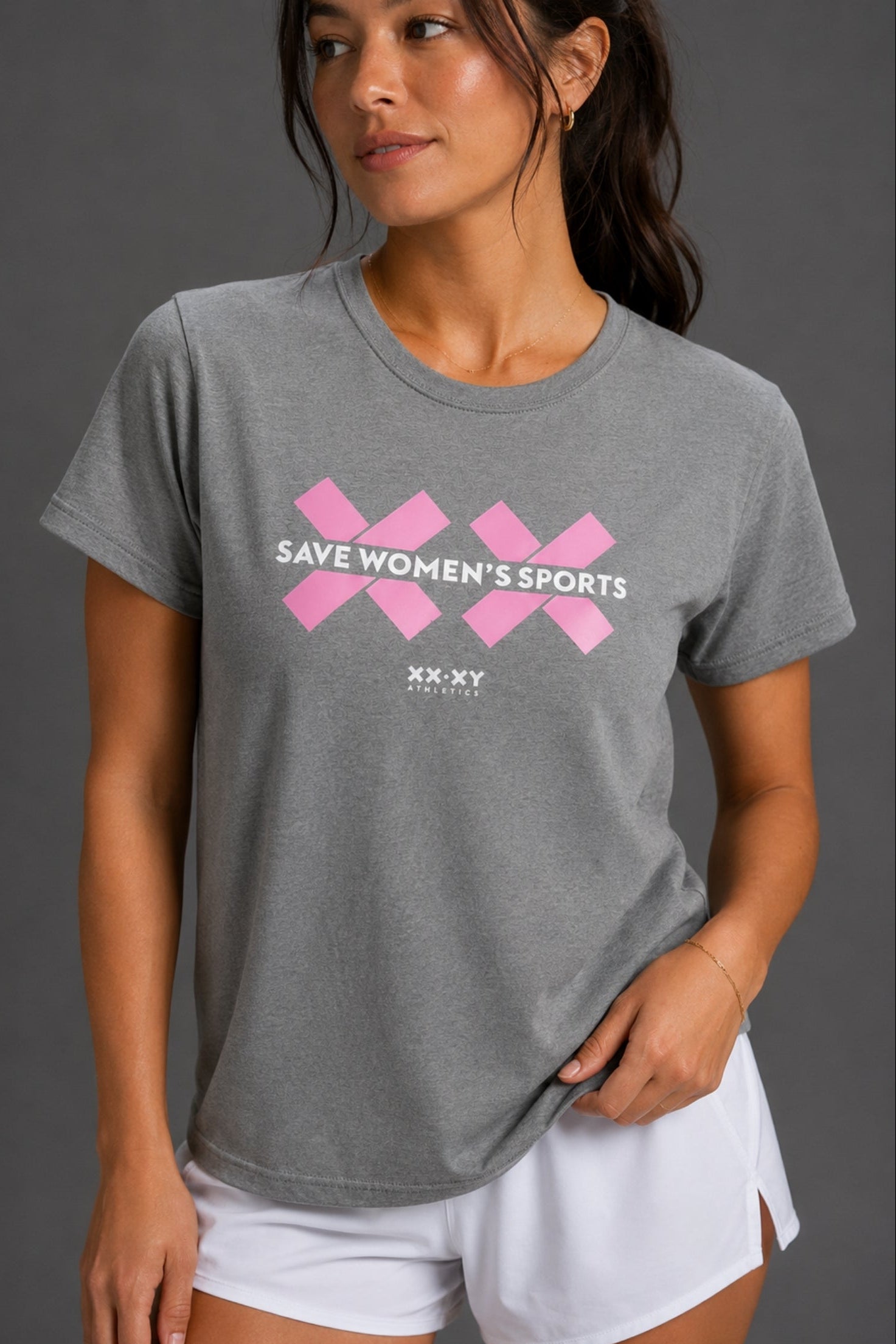 A woman wearing a heather gray crewneck t-shirt with a pink graphic that reads 'SAVE WOMEN'S SPORTS' and 'XX XY' underneath, paired with white athletic shorts against a dark gray studio background. || Heather Gray
