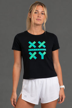 A woman modeling a black short-sleeve t-shirt with a teal and green geometric 'XXY' graphic on the chest, paired with white athletic shorts, against a solid gray studio background. || Black