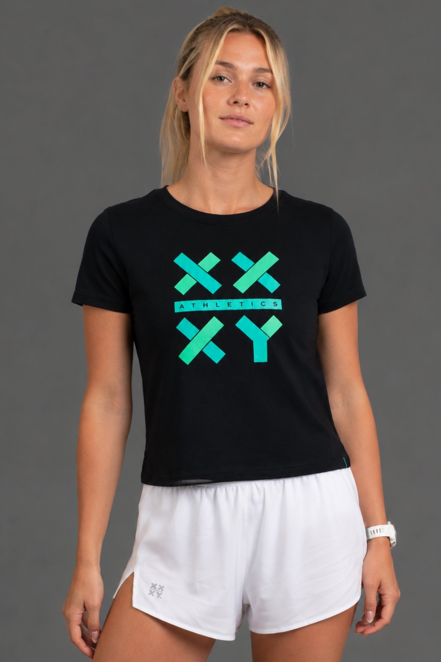 A woman modeling a black short-sleeve t-shirt with a teal and green geometric 'XXY' graphic on the chest, paired with white athletic shorts, against a solid gray studio background. || Black