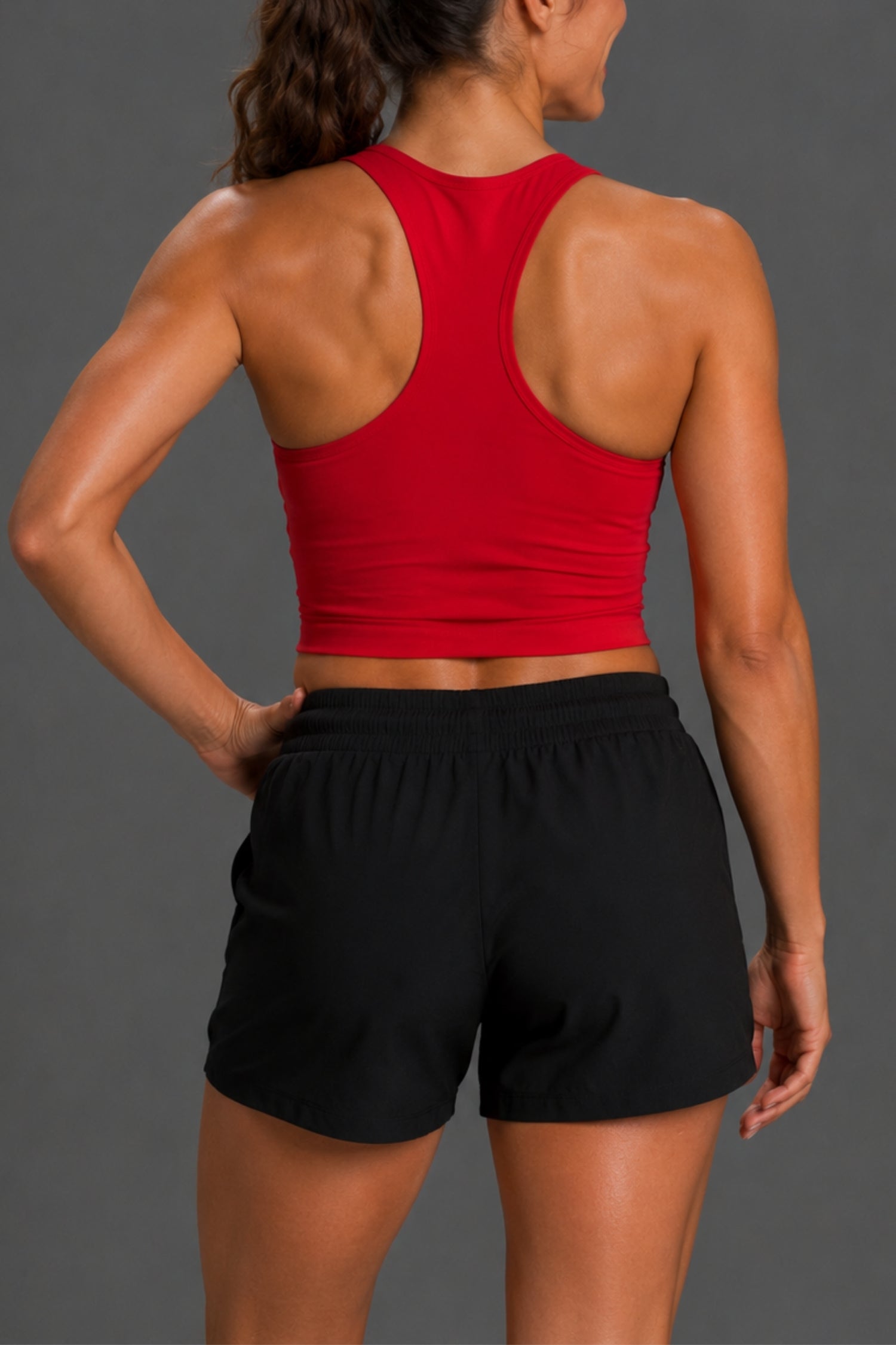 Rear view of a woman wearing a bright red racerback crop top and black athletic shorts against a dark gray studio background.
