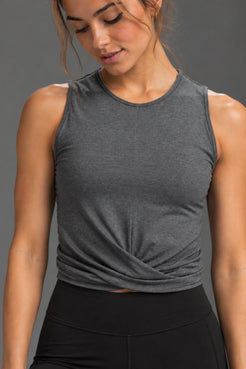 A woman wearing a dark heather gray sleeveless crop top with a twisted front detail, paired with black athletic bottoms, against a solid gray studio background. || Dark Heather Gray