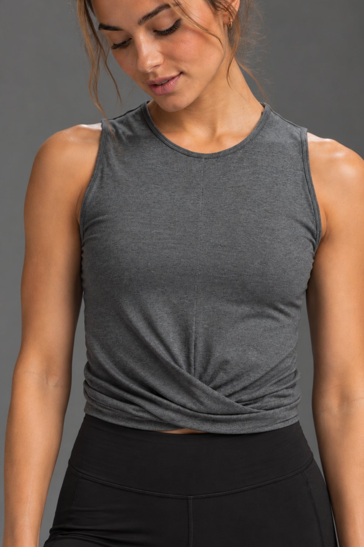 A woman wearing a dark heather gray sleeveless crop top with a twisted front detail, paired with black athletic bottoms, against a solid gray studio background. || Dark Heather Gray