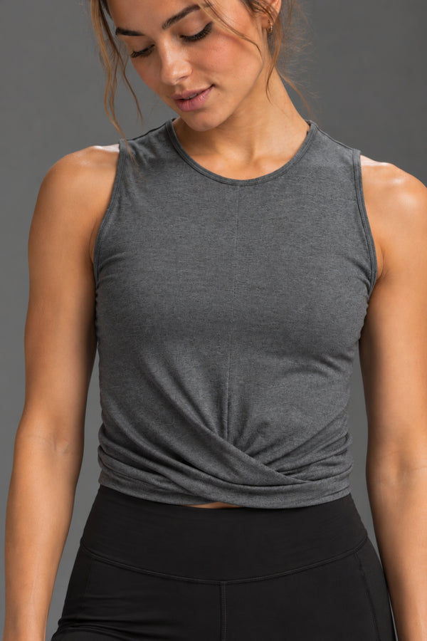 A woman wearing a dark heather gray sleeveless crop top with a twisted front detail, paired with black athletic bottoms, against a solid gray studio background. || Dark Heather Gray