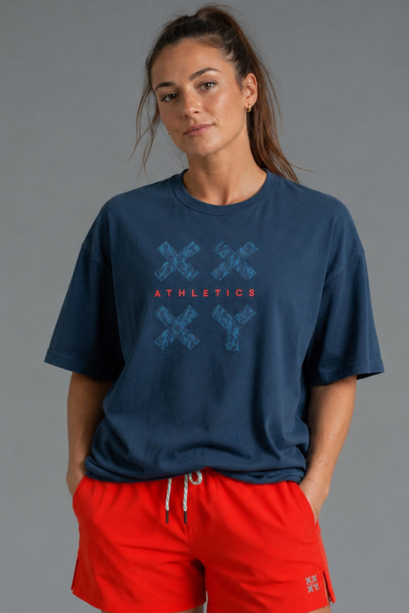 A woman wearing a navy blue t-shirt with a blue distressed graphic and the word 'ATHLETICS' in red, paired with bright red athletic shorts, against a solid gray studio background. || Navy