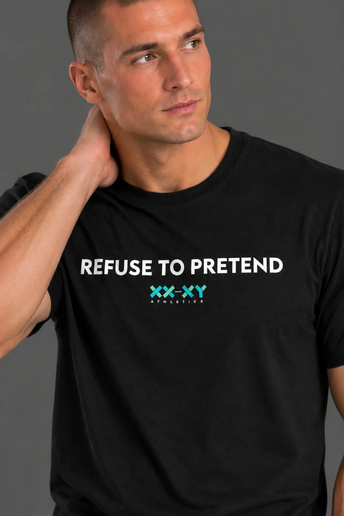 Male model wearing a black "REFUSE TO PRETEND" XX-XY Athletics T-shirt and black shorts, standing with one hand in his pocket and looking to the side.