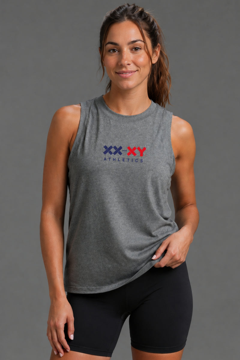 A woman modeling a heather gray sleeveless tank top with a red and navy 'XX-XY Athletics' logo on the chest, paired with black bike shorts, against a solid gray studio background. || Dark Heather Gray