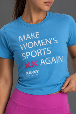 A woman wearing a bright blue athletic t-shirt with the slogan 'MAKE WOMEN'S SPORTS AGAIN' in white text, accented with pink 'XX' symbols and the 'XX-XXY ATHLETICS' logo. She is also wearing pink athletic shorts against a solid gray studio background. || Light Blue