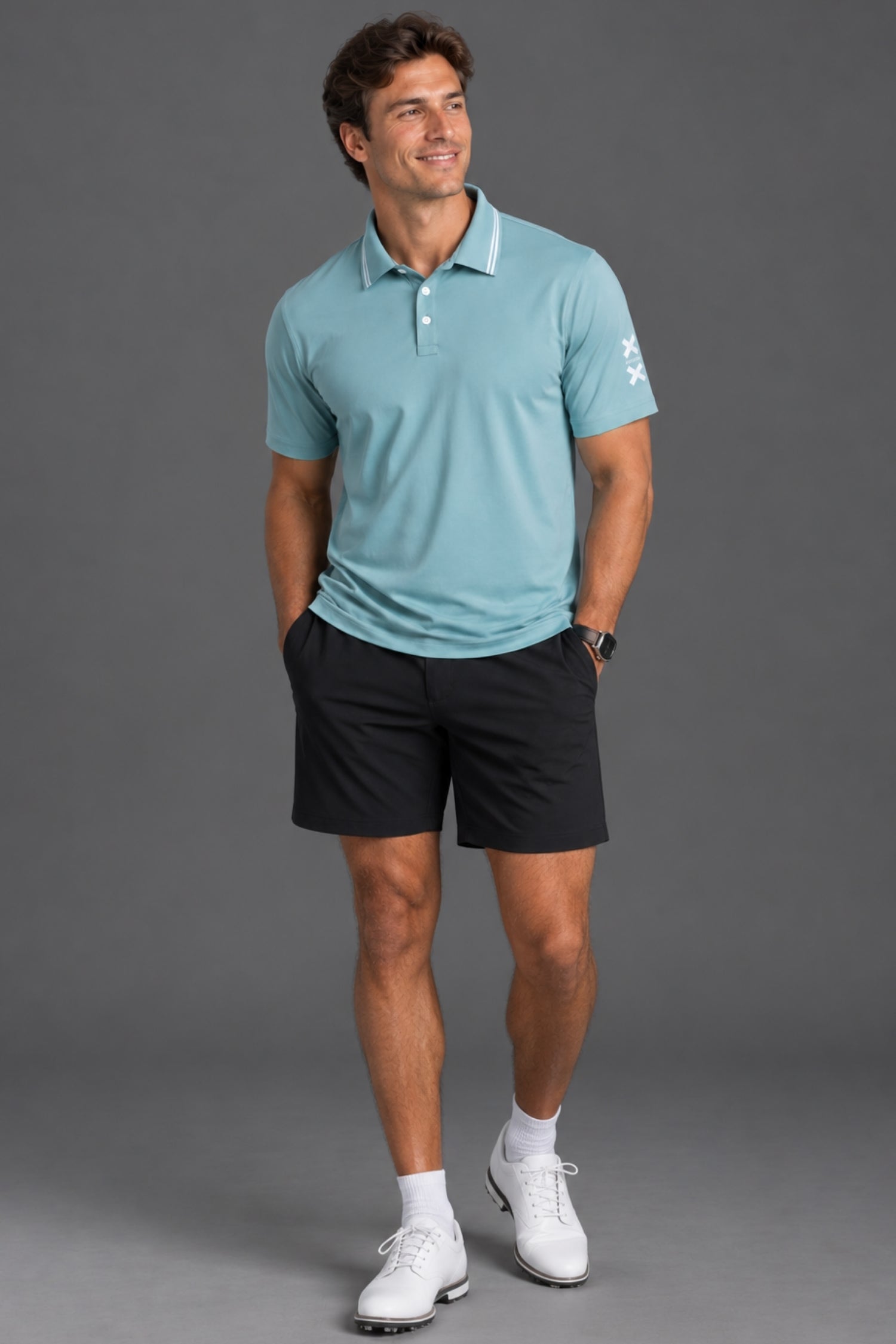 A man modeling a light blue short-sleeve polo shirt with black golf shorts, white socks, and white golf shoes against a solid gray studio background.
