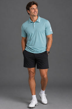 A man modeling a light blue short-sleeve polo shirt with black golf shorts, white socks, and white golf shoes against a solid gray studio background. || Underseas