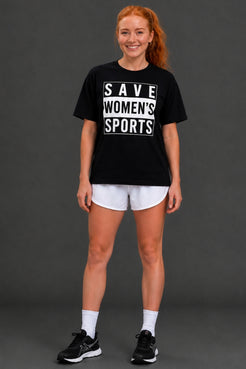 A woman modeling a black graphic t-shirt with white text reading 'SAVE WOMEN'S SPORTS', paired with white athletic shorts, white socks, and black sneakers, against a dark gray studio background. || Black