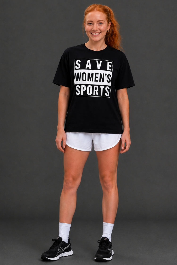 A woman modeling a black graphic t-shirt with white text reading 'SAVE WOMEN'S SPORTS', paired with white athletic shorts, white socks, and black sneakers, against a dark gray studio background. || Black