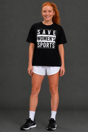 A woman modeling a black graphic t-shirt with white text reading 'SAVE WOMEN'S SPORTS', paired with white athletic shorts, white socks, and black sneakers, against a dark gray studio background. || Black