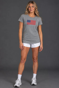 Female model wearing a gray XX-XY Athletics patriotic graphic tee, white athletic shorts, white crew socks, and sneakers, standing against a gray studio background || Heather Gray