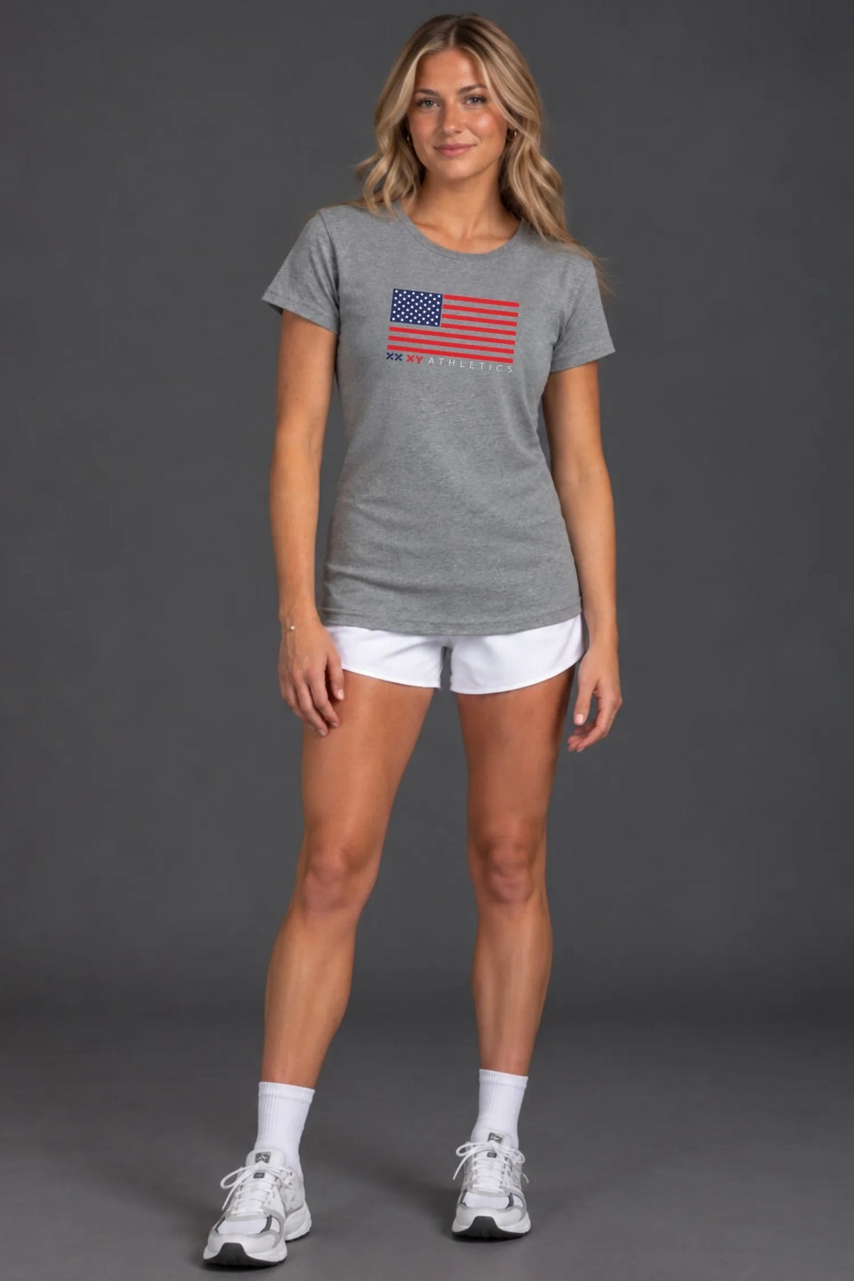 Female model wearing a gray XX-XY Athletics patriotic graphic tee, white athletic shorts, white crew socks, and sneakers, standing against a gray studio background || Heather Gray