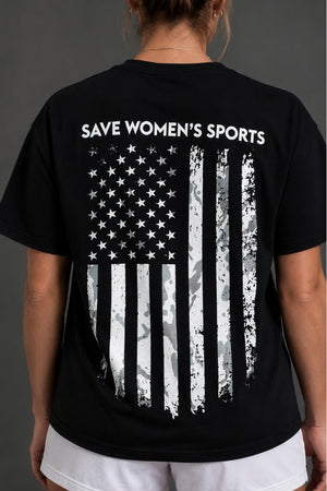 A person viewed from the back wearing a black short-sleeve T-shirt with the slogan 'SAVE WOMEN'S SPORTS' in white above a distressed American flag graphic in black and white. || Black