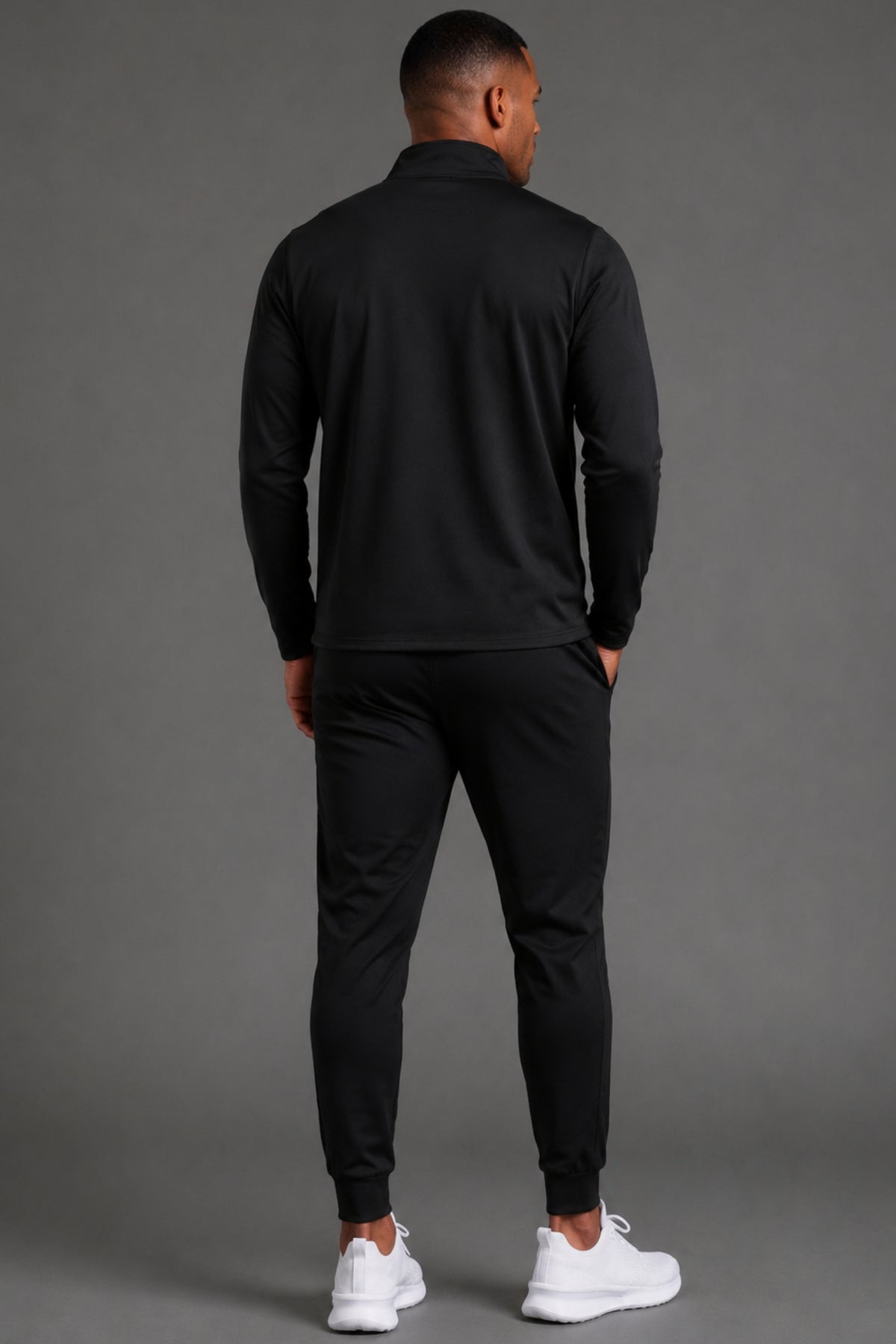 Man standing facing backwards while wearing black performance joggers styled casually with a black top and sneakers in a studio setting.