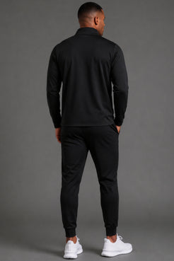 Man standing facing backwards while wearing black performance joggers styled casually with a black top and sneakers in a studio setting.