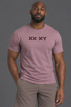 A muscular man wearing a heathered dusty rose athletic t-shirt with a dark maroon 'XX-XY ATHLETICS' logo on the chest, paired with taupe shorts, against a solid dark gray studio background. || Burgundy Heather