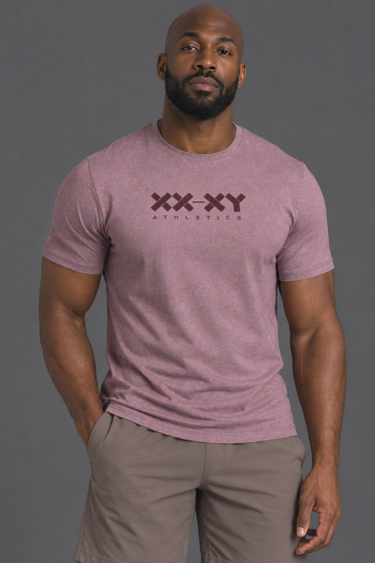 A muscular man wearing a heathered dusty rose athletic t-shirt with a dark maroon 'XX-XY ATHLETICS' logo on the chest, paired with taupe shorts, against a solid dark gray studio background. || Burgundy Heather