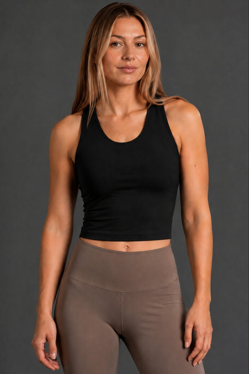 A woman modeling a black sleeveless scoop-neck athletic crop top paired with high-waisted taupe leggings, set against a solid dark gray studio background. || Black