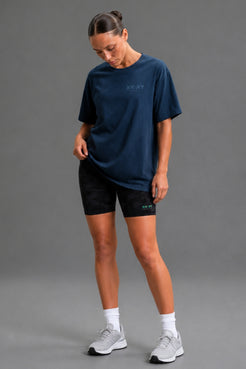 A woman modeling an oversized navy blue t-shirt with a subtle tonal logo on the chest, paired with black athletic shorts, white socks, and light gray running shoes against a solid gray studio background. || Navy