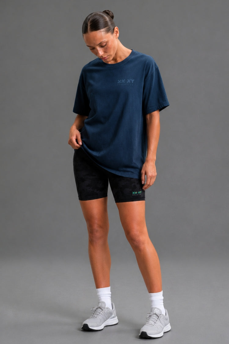 A woman modeling an oversized navy blue t-shirt with a subtle tonal logo on the chest, paired with black athletic shorts, white socks, and light gray running shoes against a solid gray studio background. || Navy