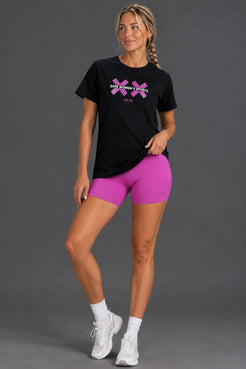 A woman modeling a black short-sleeve t-shirt with a pink graphic logo and bright pink athletic biker shorts, paired with white socks and white sneakers, against a solid gray studio background. || Bright Fuchsia