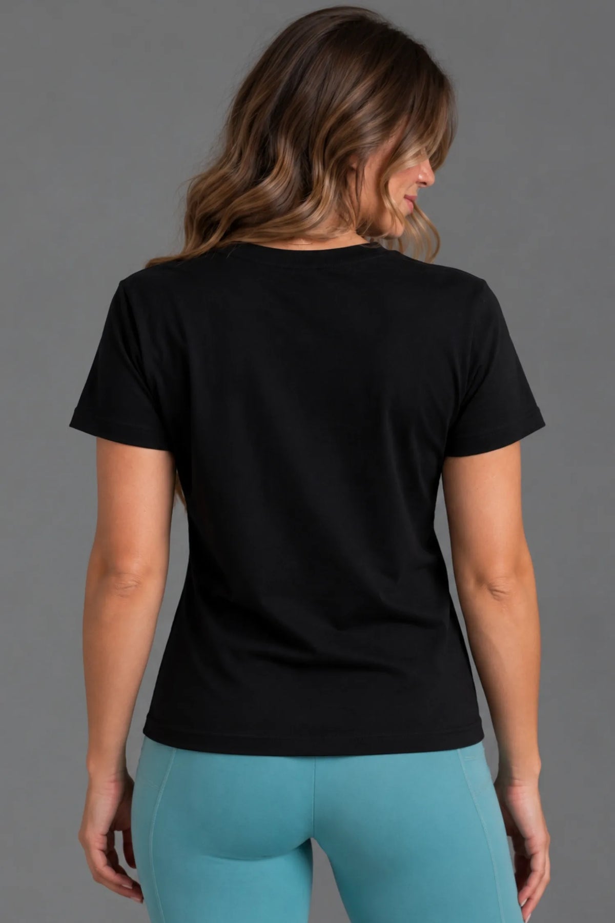 Back view of a female model wearing a blank black tee with teal leggings. || Black