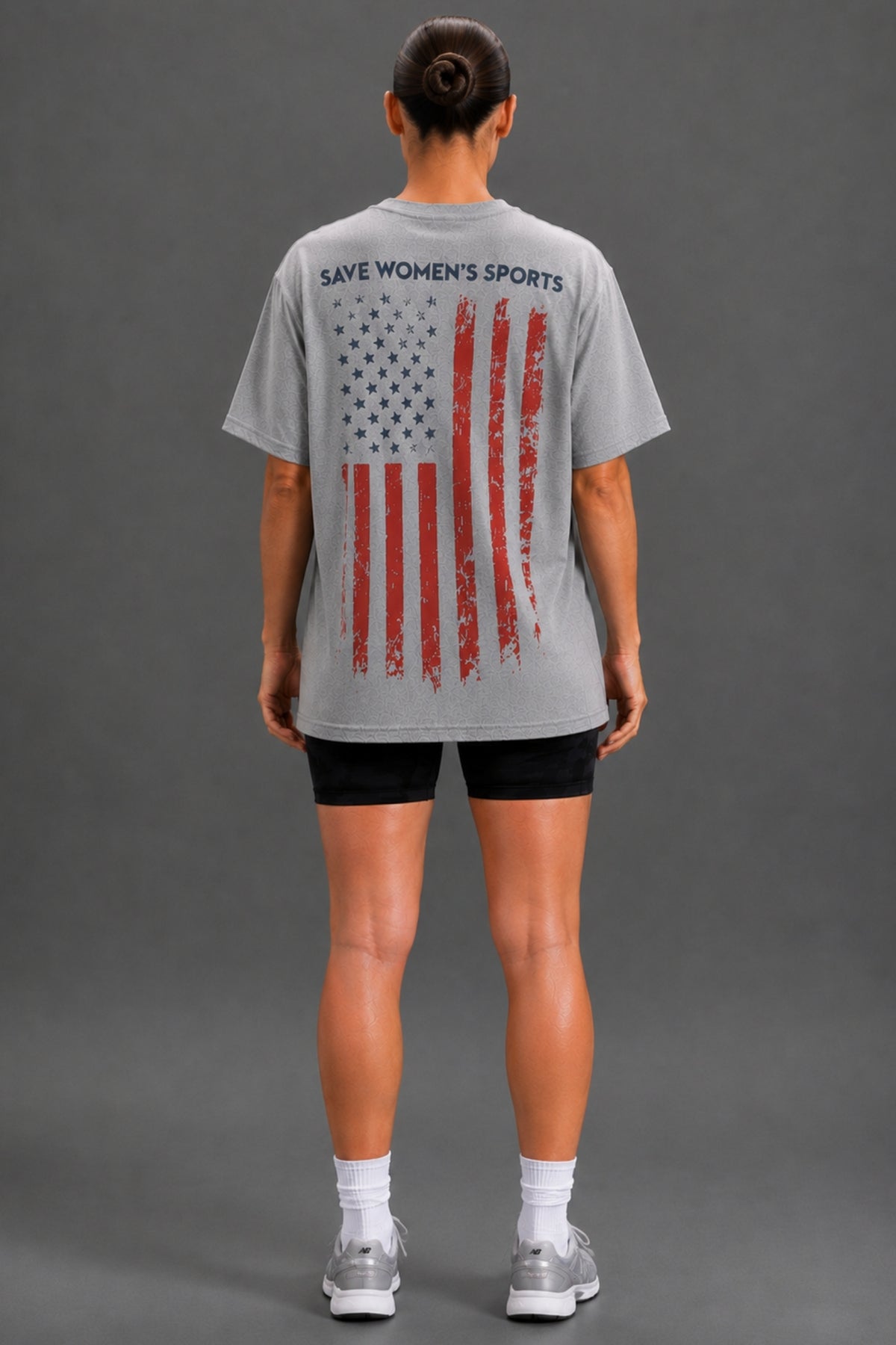 A woman viewed from the back wearing a light gray oversized T-shirt with a distressed American flag graphic and the text 'SAVE WOMEN'S SPORTS' across the upper back, paired with black athletic shorts, white socks, and gray sneakers against a dark gray studio background.