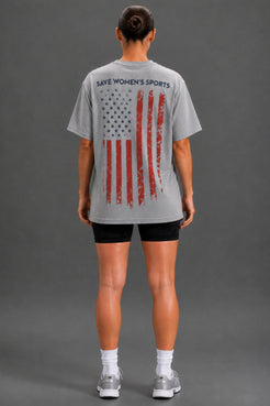 A woman viewed from the back wearing a light gray oversized T-shirt with a distressed American flag graphic and the text 'SAVE WOMEN'S SPORTS' across the upper back, paired with black athletic shorts, white socks, and gray sneakers against a dark gray studio background. || Heather Gray