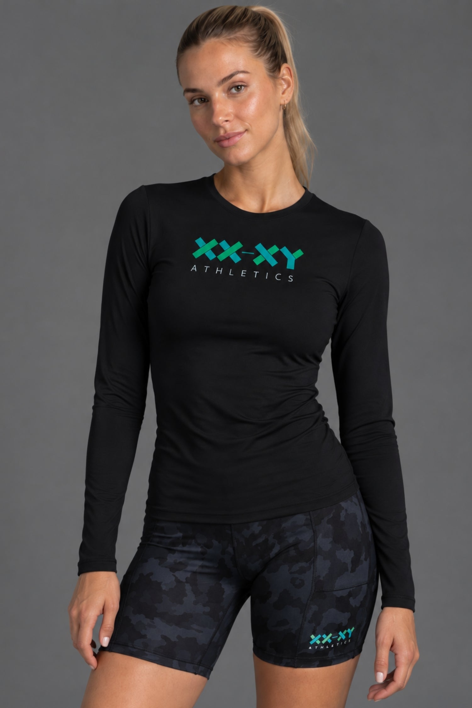A woman modeling a black long-sleeve athletic top with a teal and white 'XX-XY Athletics' logo on the chest, paired with dark camouflage athletic shorts against a solid gray studio background. || Black