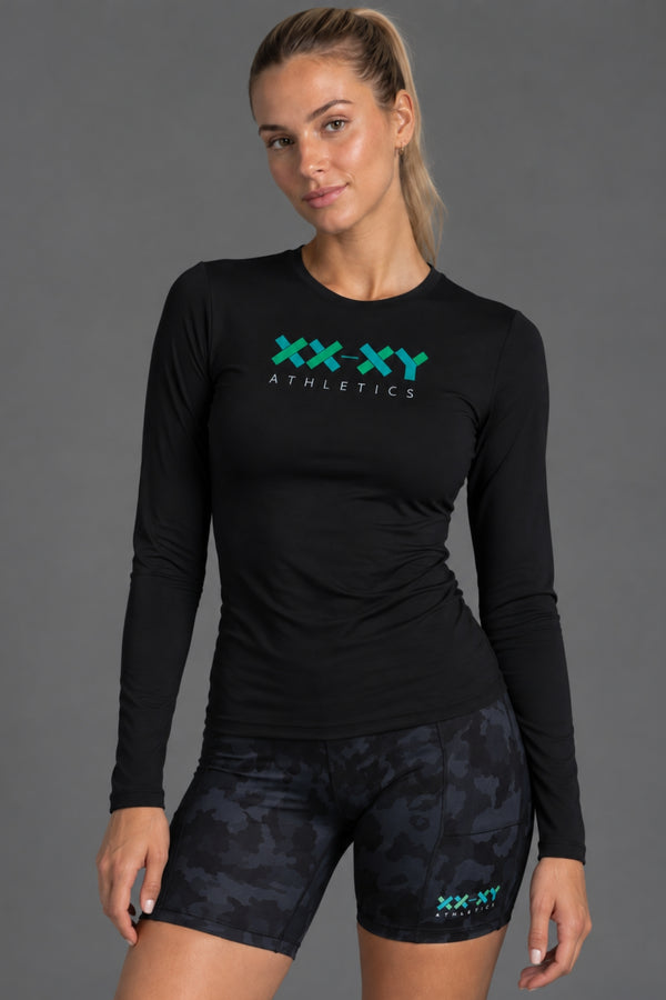 A woman modeling a black long-sleeve athletic top with a teal and white 'XX-XY Athletics' logo on the chest, paired with dark camouflage athletic shorts against a solid gray studio background. || Black