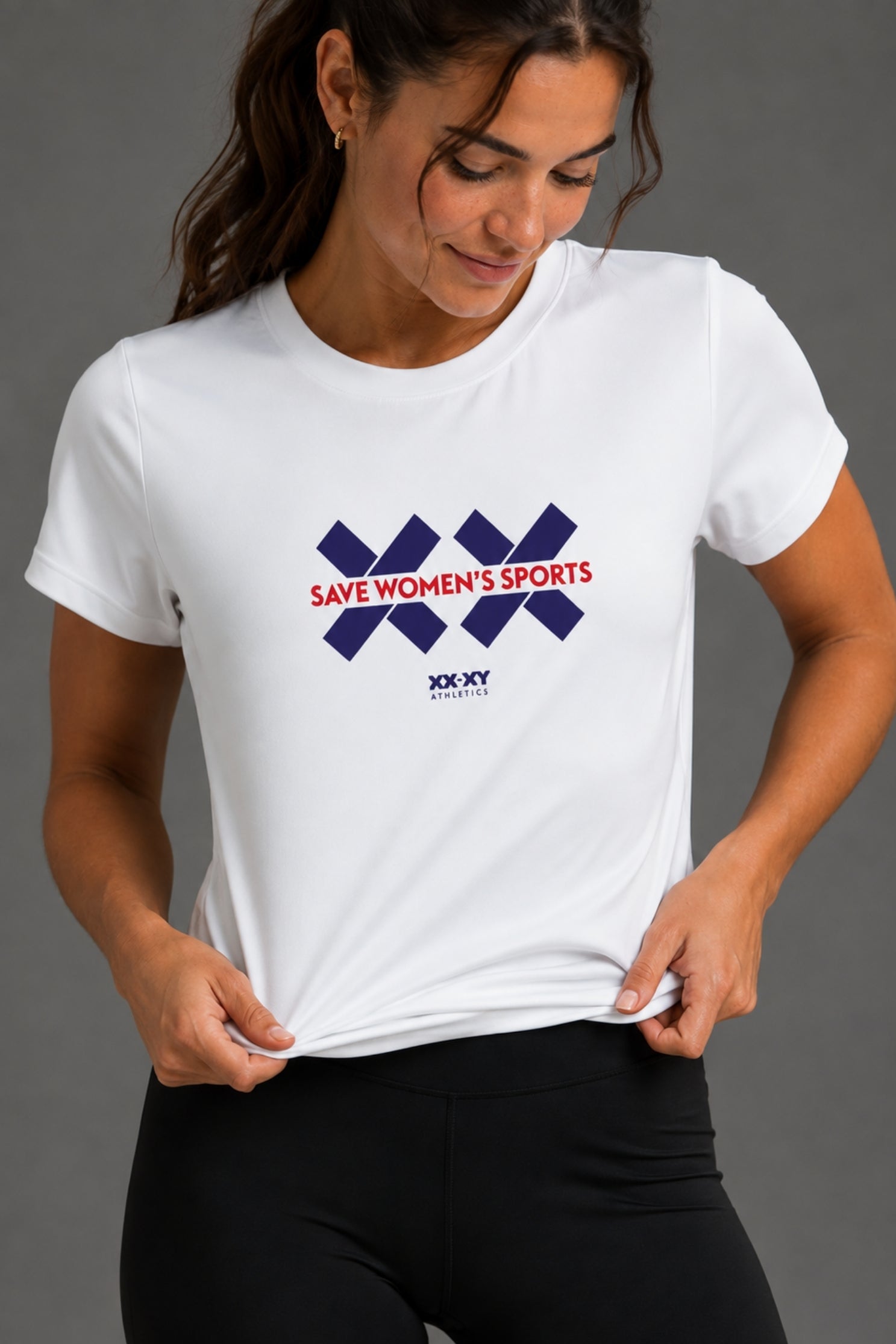 A woman wearing a white short-sleeve athletic T-shirt with a navy and red 'SAVE WOMEN'S SPORTS' graphic on the chest, paired with black leggings, against a dark gray studio background. || White