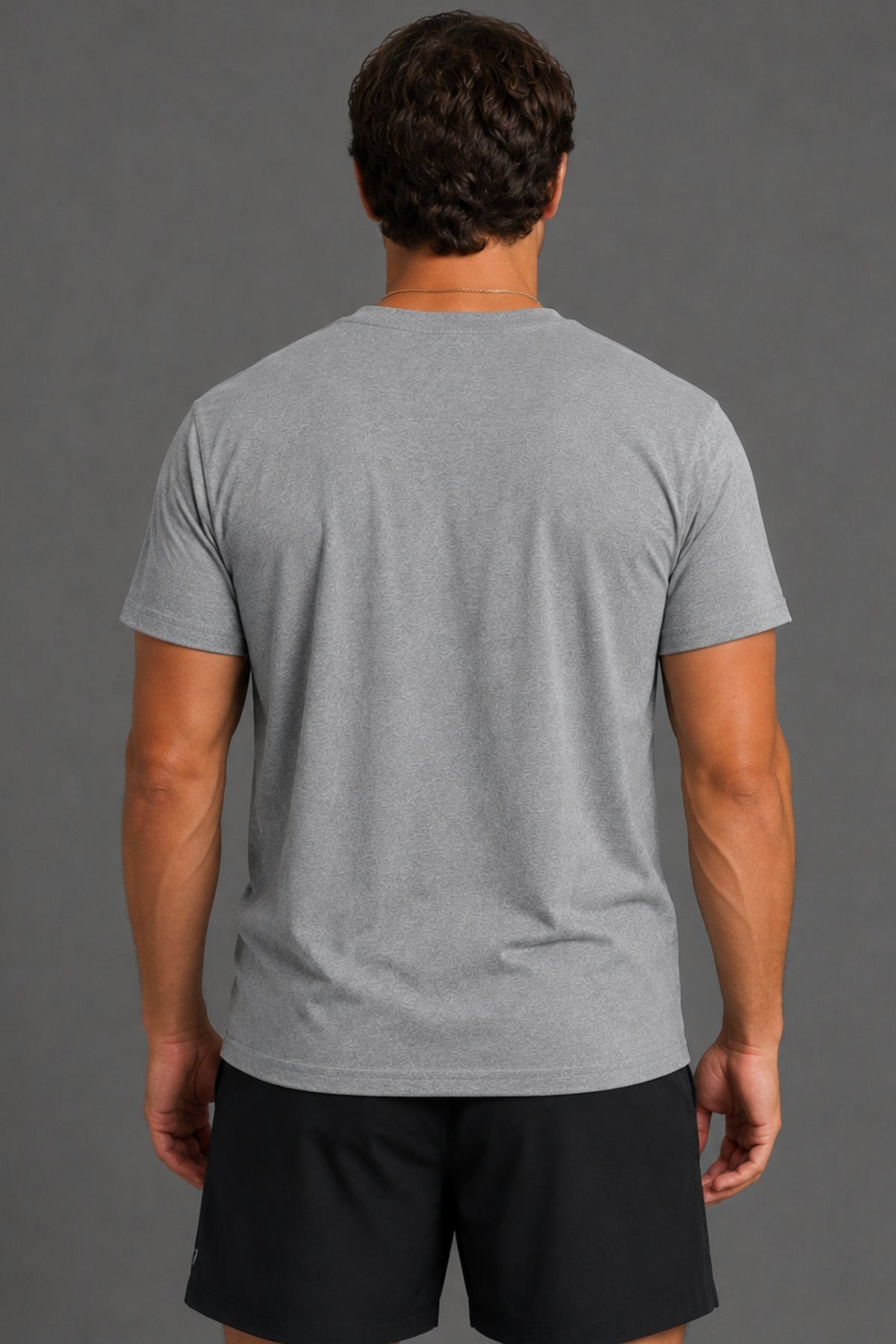 A man viewed from behind wearing a heather gray short-sleeve t-shirt and black athletic shorts against a solid gray studio background.