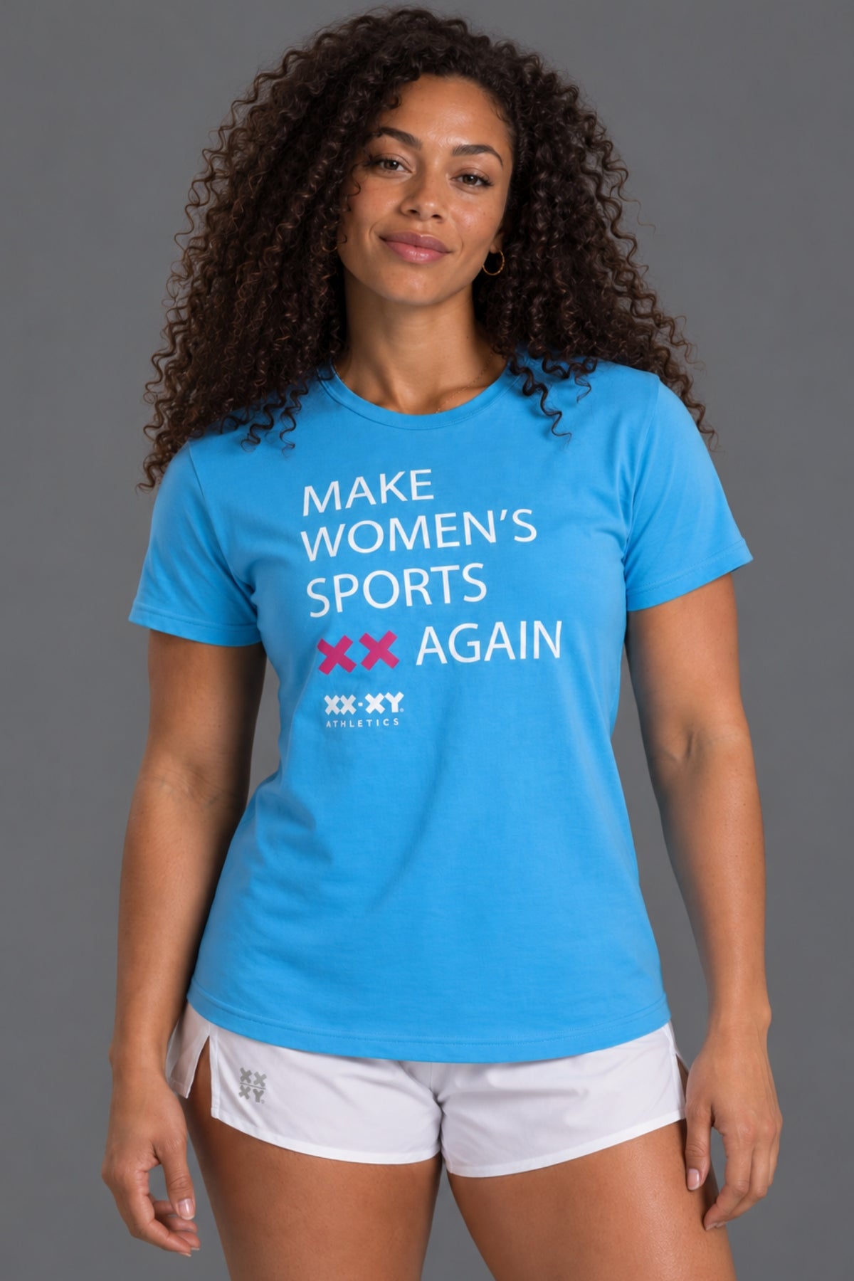 A woman modeling a bright blue short-sleeve graphic T-shirt with the text 'MAKE WOMEN'S SPORTS XX AGAIN' and 'XX-XY ATHLETICS' printed on the front, paired with white athletic shorts against a solid gray studio background.