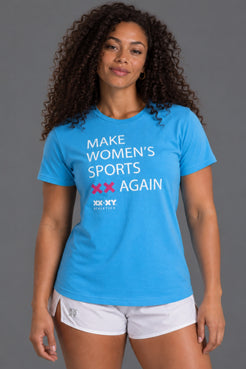A woman modeling a bright blue short-sleeve graphic T-shirt with the text 'MAKE WOMEN'S SPORTS XX AGAIN' and 'XX-XY ATHLETICS' printed on the front, paired with white athletic shorts against a solid gray studio background. || Light Blue