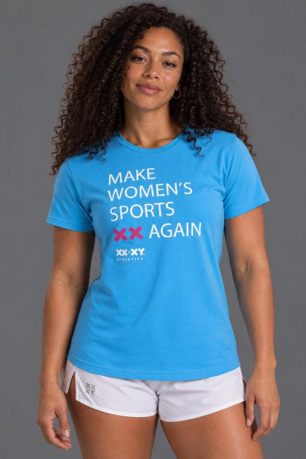 A woman modeling a bright blue short-sleeve graphic T-shirt with the text 'MAKE WOMEN'S SPORTS XX AGAIN' and 'XX-XY ATHLETICS' printed on the front, paired with white athletic shorts against a solid gray studio background. || Light Blue