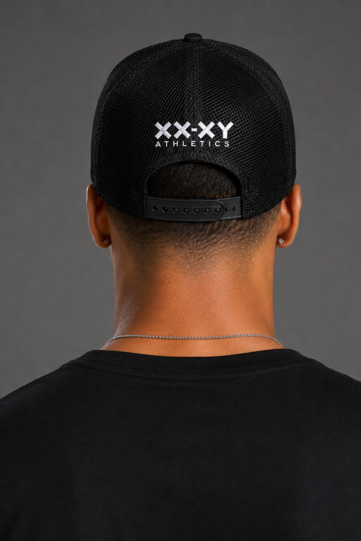 Back view of a man wearing a black mesh trucker cap with 'XX-XY ATHLETICS' printed in white on the back, set against a dark gray studio background.
