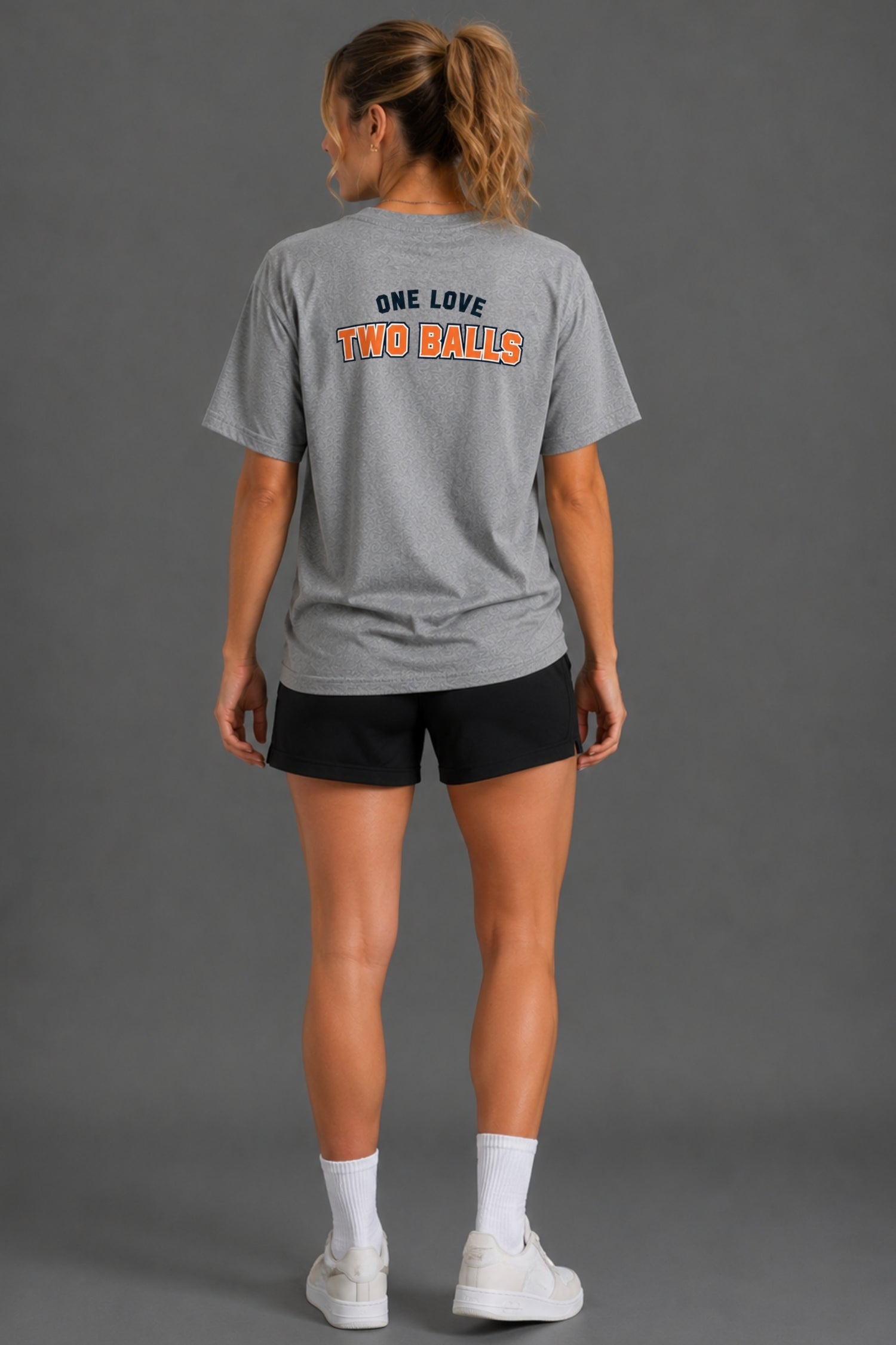 A woman shown from the back wearing a heather gray t-shirt with 'ONE LOVE TWO BALLS' printed in navy and orange on the back, paired with black shorts, white socks, and white sneakers against a solid gray studio background.