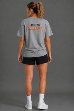 A woman shown from the back wearing a heather gray t-shirt with 'ONE LOVE TWO BALLS' printed in navy and orange on the back, paired with black shorts, white socks, and white sneakers against a solid gray studio background. || Heather Gray