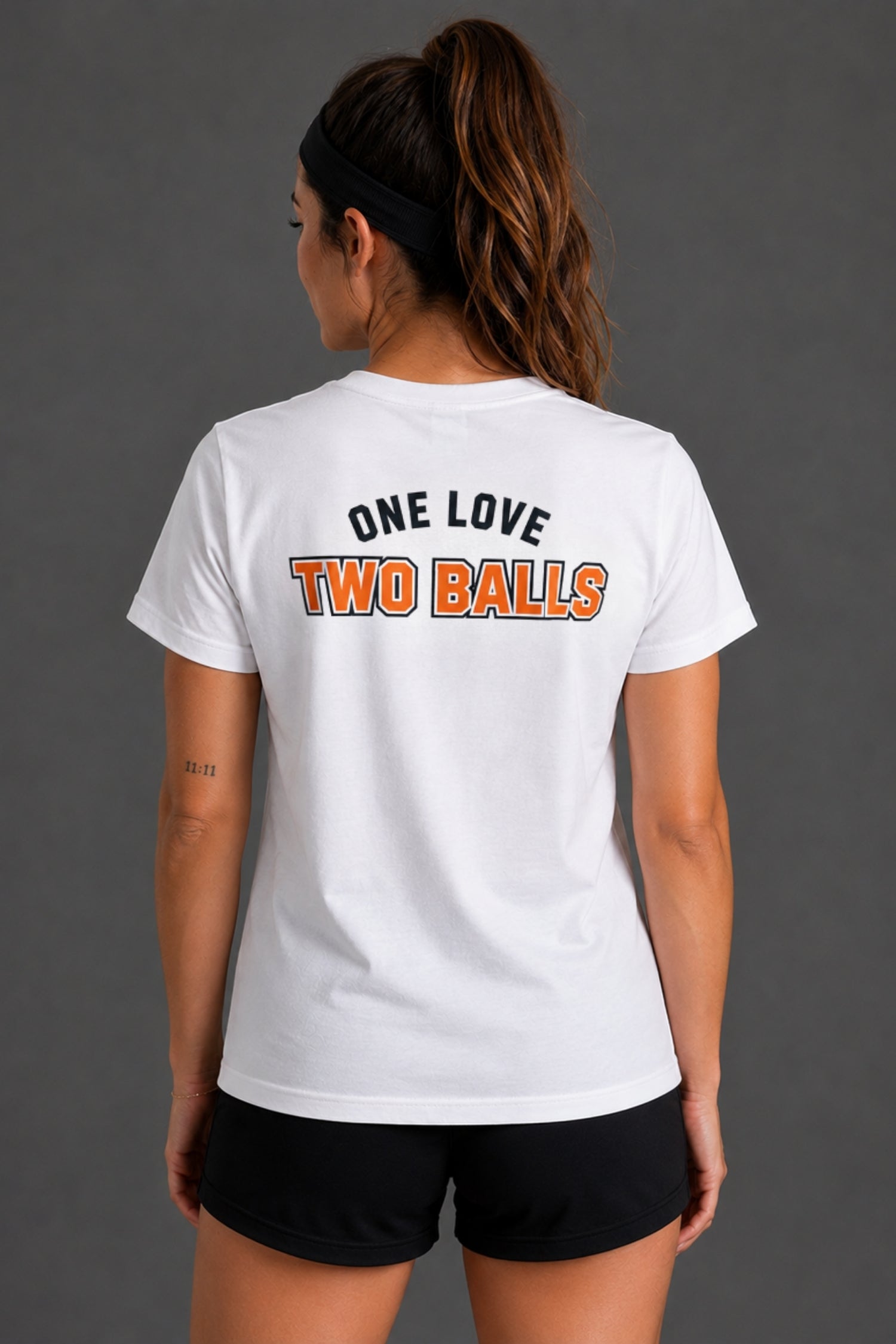 A woman shown from the back wearing a white short-sleeve graphic T-shirt with the text 'ONE LOVE TWO BALLS' printed across the upper back in black and orange, paired with black athletic shorts against a dark gray studio background.