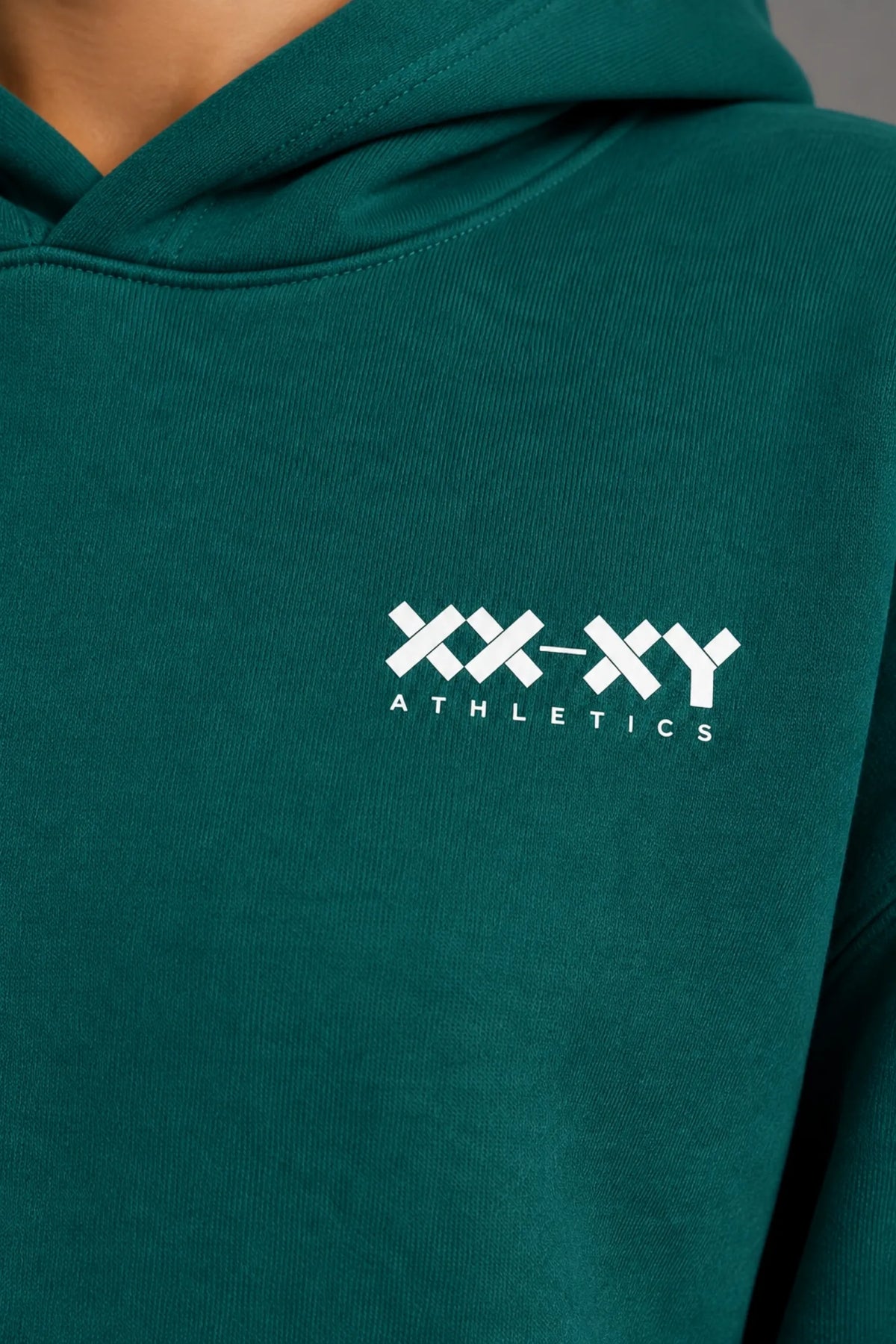 Close-up of the white XX-XY Athletics logo on the chest of a teal hoodie. || Pine