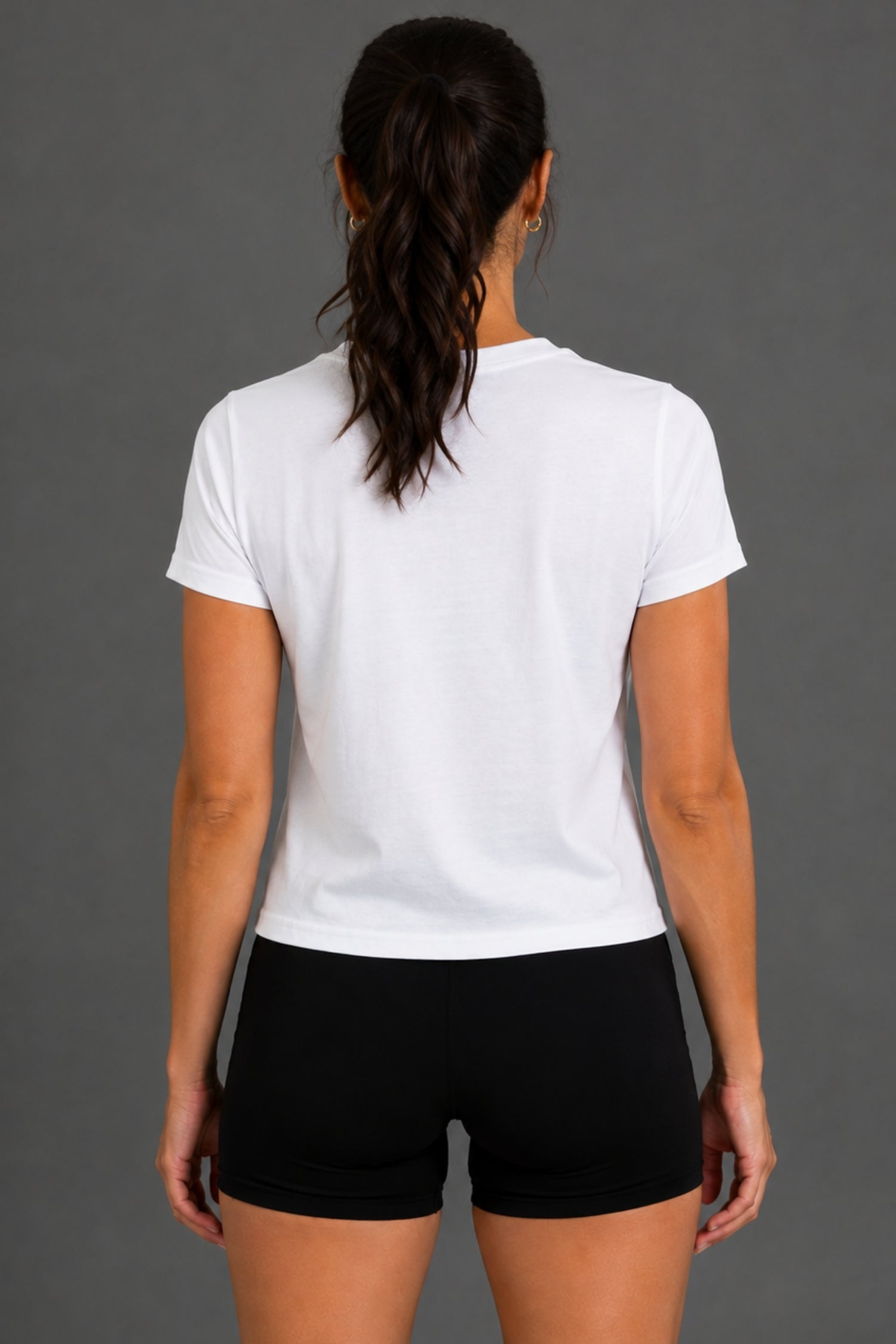 A woman viewed from the back wearing a plain white short-sleeve crewneck T-shirt and black athletic shorts against a solid gray studio background. || White
