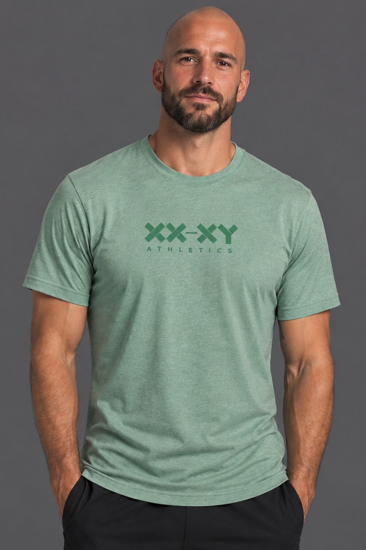 A man wearing a heather green crewneck t-shirt with a dark green 'XX-XY ATHLETICS' logo printed on the chest, posed against a solid dark gray studio background.