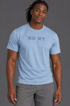 A man wearing a light blue crewneck t-shirt with a dark gray 'XX-XY ATHLETICS' logo on the chest, paired with gray athletic pants, against a solid gray studio background. || Cerulean Blue Heather