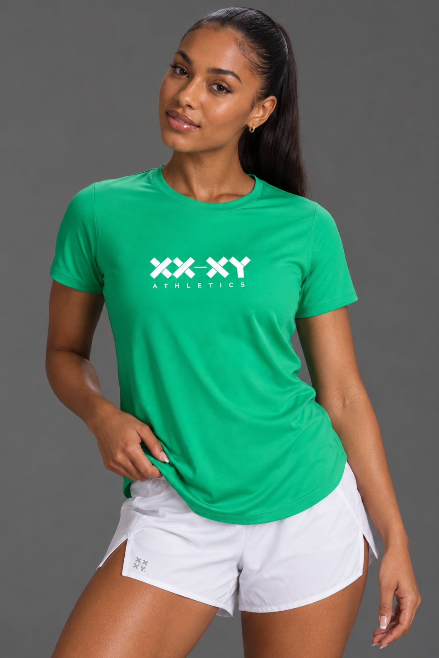 Female model wearing a green XX-XY Athletics T-shirt and white running shorts, posed against a gray studio background. || Kelly Green