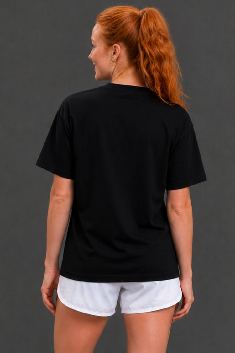 A woman with red hair in a ponytail is shown from the back, wearing a plain black short-sleeve t-shirt and white athletic shorts against a dark gray studio background.