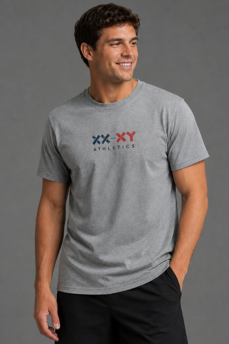 A man wearing a heather gray crewneck t-shirt with a red and blue 'XX XY ATHLETICS' logo on the chest, paired with black pants, set against a solid gray studio background. || Heather Gray