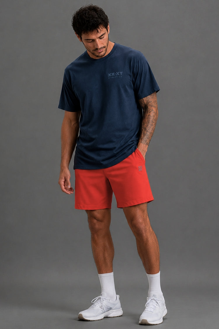 A man viewed from the front wearing a navy blue t-shirt with a XX_XY ATHLETICS graphic on the chest, paired with red athletic shorts, white socks, and gray sneakers, against a solid gray studio background.