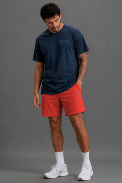 A man viewed from the front wearing a navy blue t-shirt with a XX_XY ATHLETICS graphic on the chest, paired with red athletic shorts, white socks, and gray sneakers, against a solid gray studio background. || Navy
